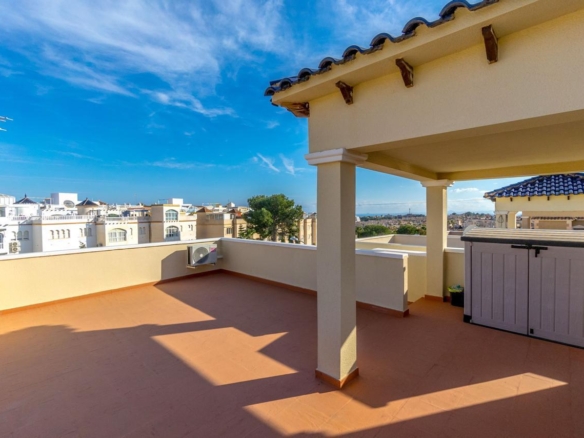 Sunny rooftop terrace with terracotta floor, beige columns, and a gray storage shed, overlooking a distant cityscape under a blue sky.