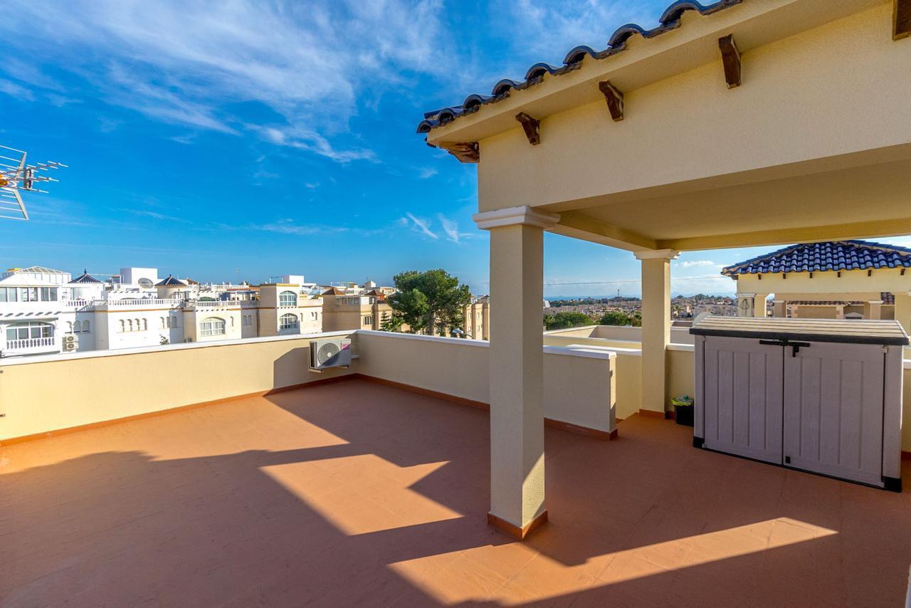 Sunny rooftop terrace with terracotta floor, beige columns, and a gray storage shed, overlooking a distant cityscape under a blue sky.