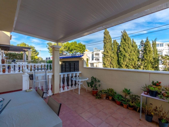 Sunlit terrace with terracotta tiles, white railing, and a row of potted plants along the wall beside a table covered with a cloth and chairs nearby.