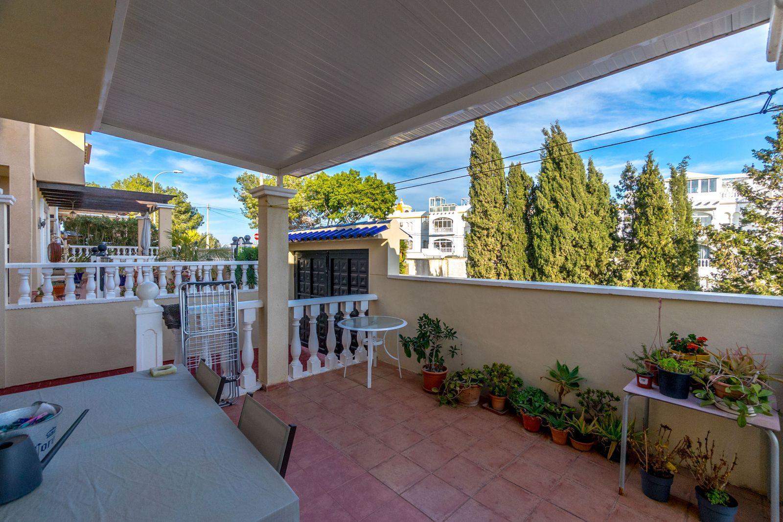 Sunlit terrace with terracotta tiles, white railing, and a row of potted plants along the wall beside a table covered with a cloth and chairs nearby.