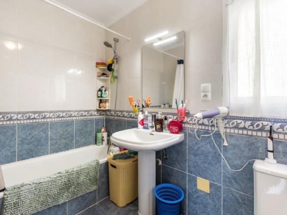 Small bathroom with a pedestal sink, mirror, and bathtub; toiletries line the tub edge and a towel hangs over the tub side.