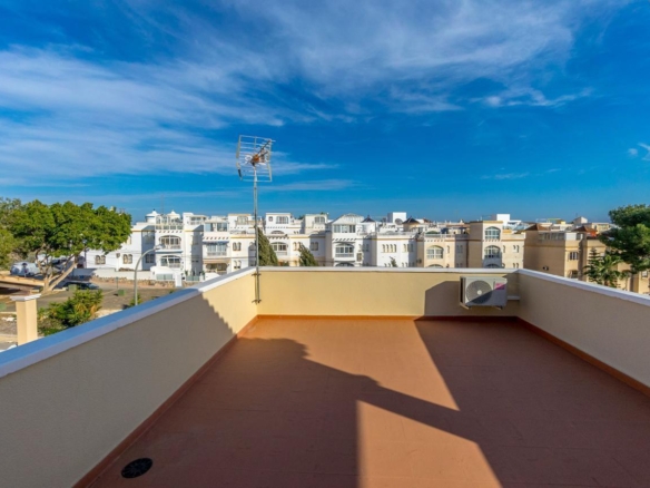Rooftop terrace with brown tiles, a satellite antenna, and a wall-mounted AC unit, overlooking white apartment buildings under a blue sky.