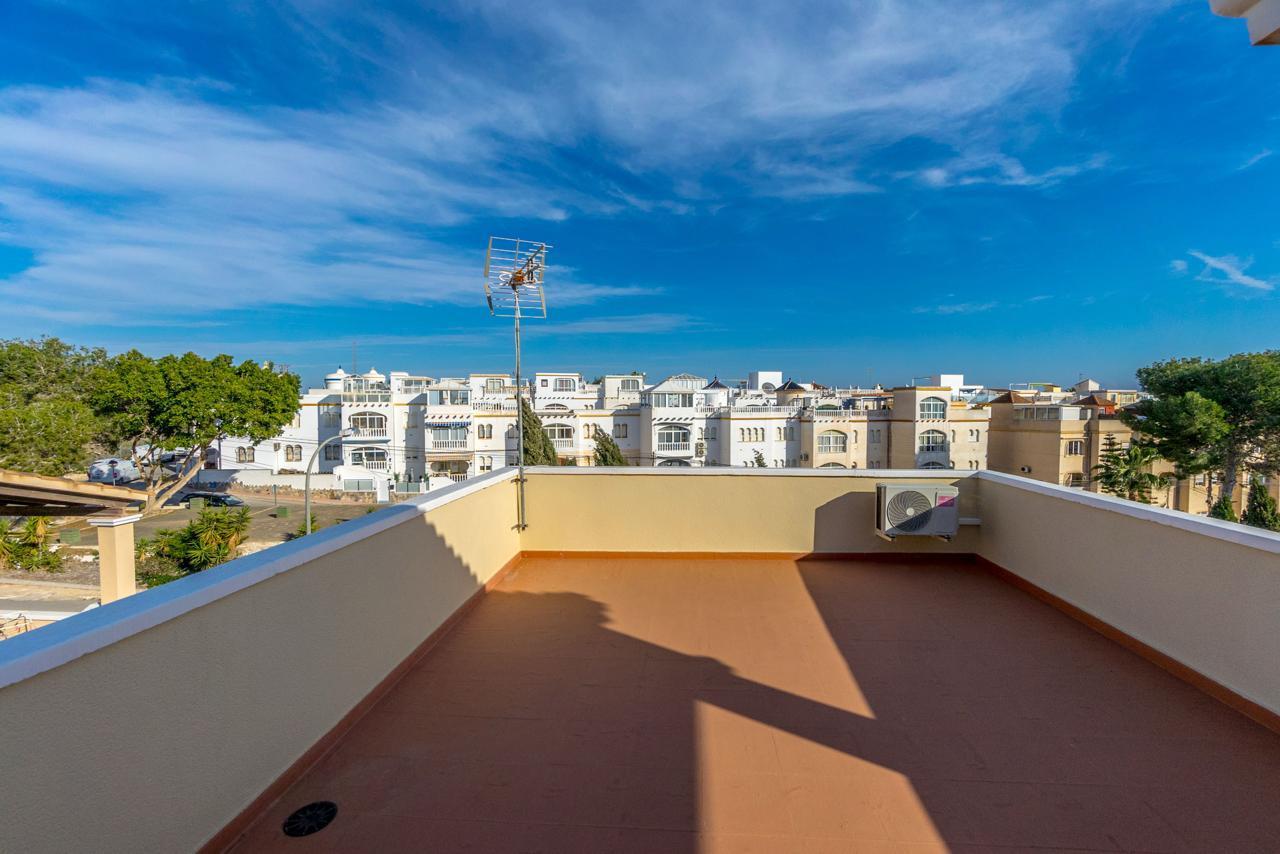Rooftop terrace with brown tiles, a satellite antenna, and a wall-mounted AC unit, overlooking white apartment buildings under a blue sky.