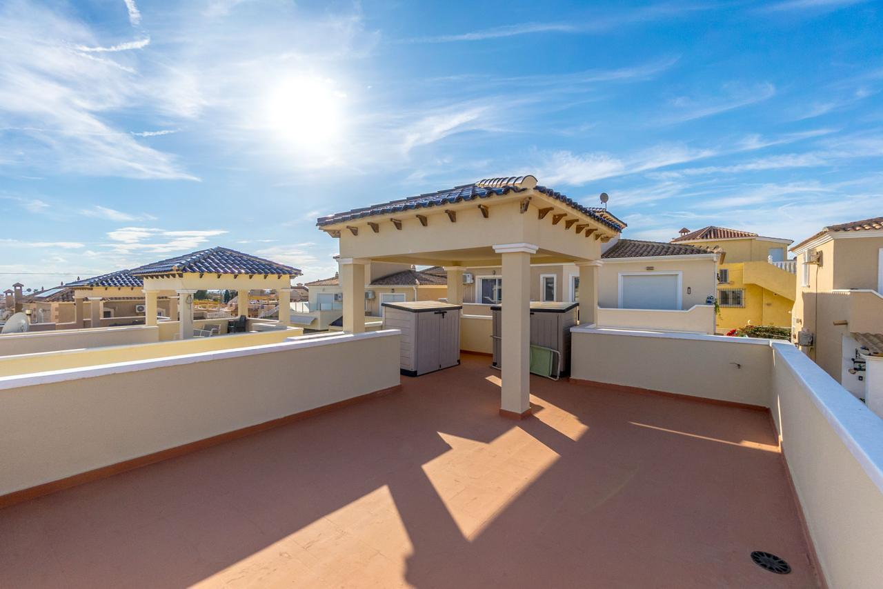 Sunlit rooftop terrace with beige walls, terra-cotta floor, and two small tiled pergolas overlooking neighboring houses.