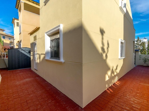 Sunny courtyard beside a beige-yellow building with a red tile floor and white-trimmed window bars.