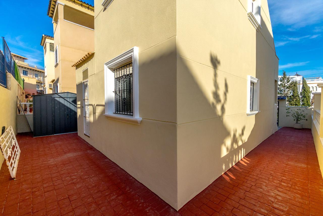 Sunny courtyard beside a beige-yellow building with a red tile floor and white-trimmed window bars.