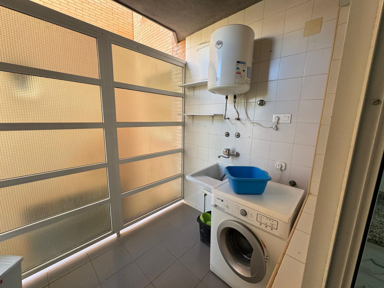 Laundry room with front-loading washing machine, blue tub, and sink under a wall-mounted water heater; shelves above the sink and a large frosted glass door on the left.