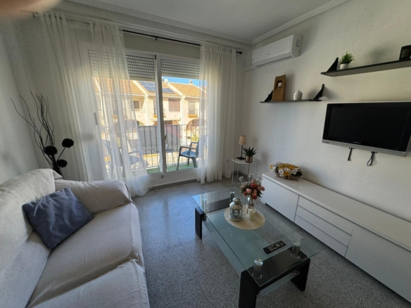 Bright living room with a white sofa, glass coffee table, and wall-mounted TV facing a balcony door with sheer white curtains.