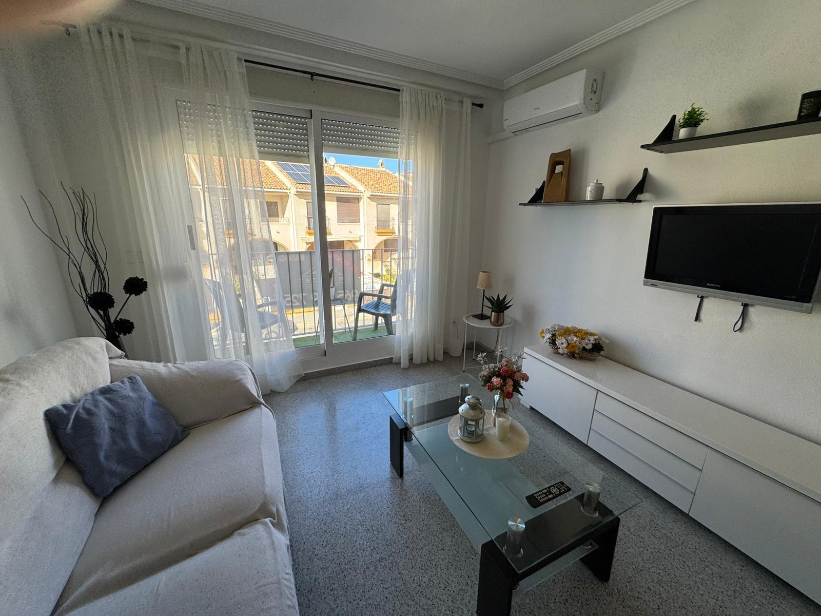 Bright living room with a white sofa, glass coffee table, and wall-mounted TV facing a balcony door with sheer white curtains.