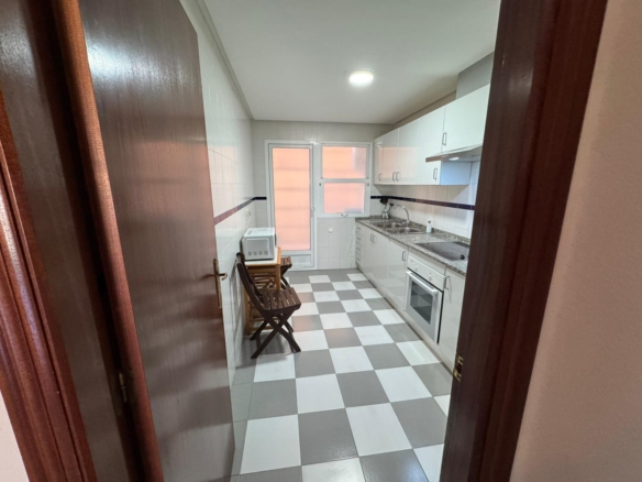 Narrow white kitchen viewed from doorway: checkered gray and white floor, white cabinets, sink, stove, oven, and a microwave on a small table with two wooden folding chairs nearby.