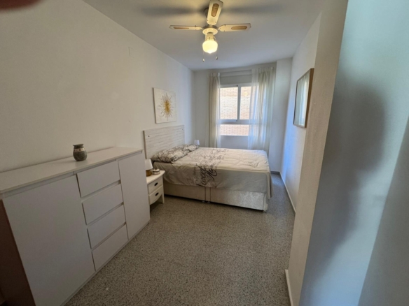 Small white bedroom with a double bed, dresser, and nightstand, near a window with sheer curtains and a sunburst wall art above the headboard.