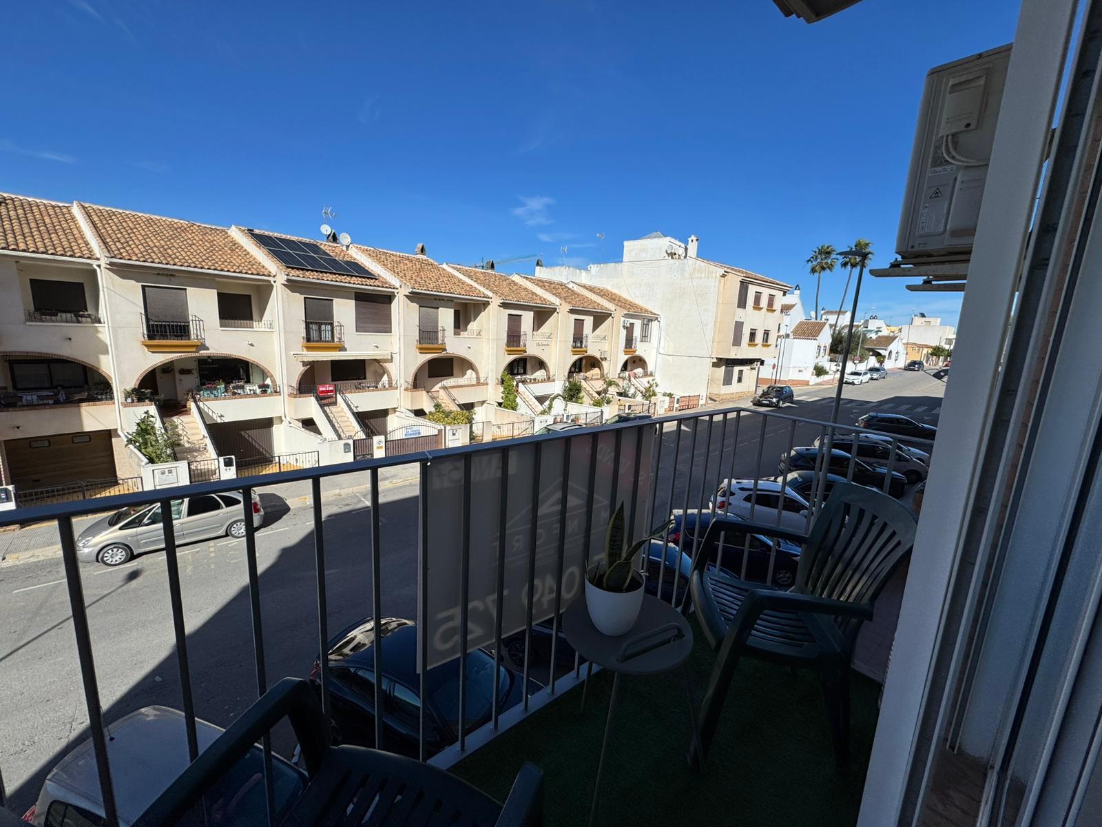 View from a balcony overlooking a sunny street lined with beige townhouses, arches on the ground floor, parked cars, and palm trees under a clear blue sky.