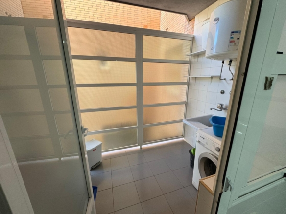Laundry area with a front-loading washing machine, white sink with blue bucket, wall-mounted water heater, shelves, and frosted glass partitioned windows.
