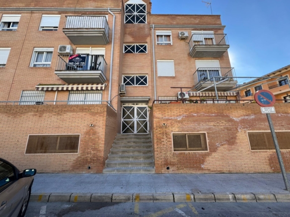 Brick apartment building with central concrete stairs and metal gate at the entrance; multiple balconies and a no-parking sign on the street corner.