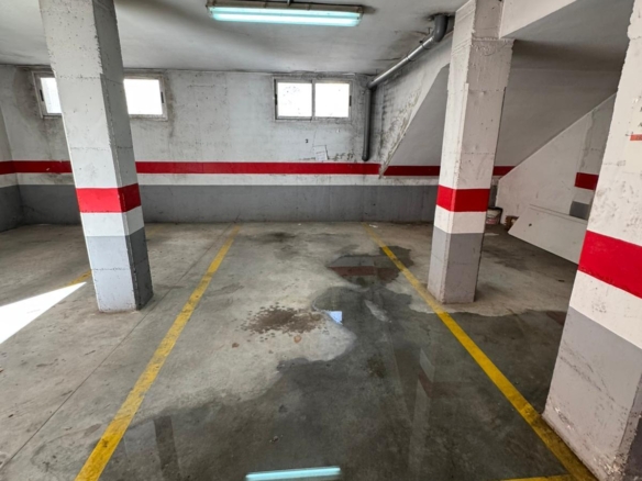 Underground parking garage with concrete pillars, yellow parking lines, and wet patches on the floor under a fluorescent ceiling light.
