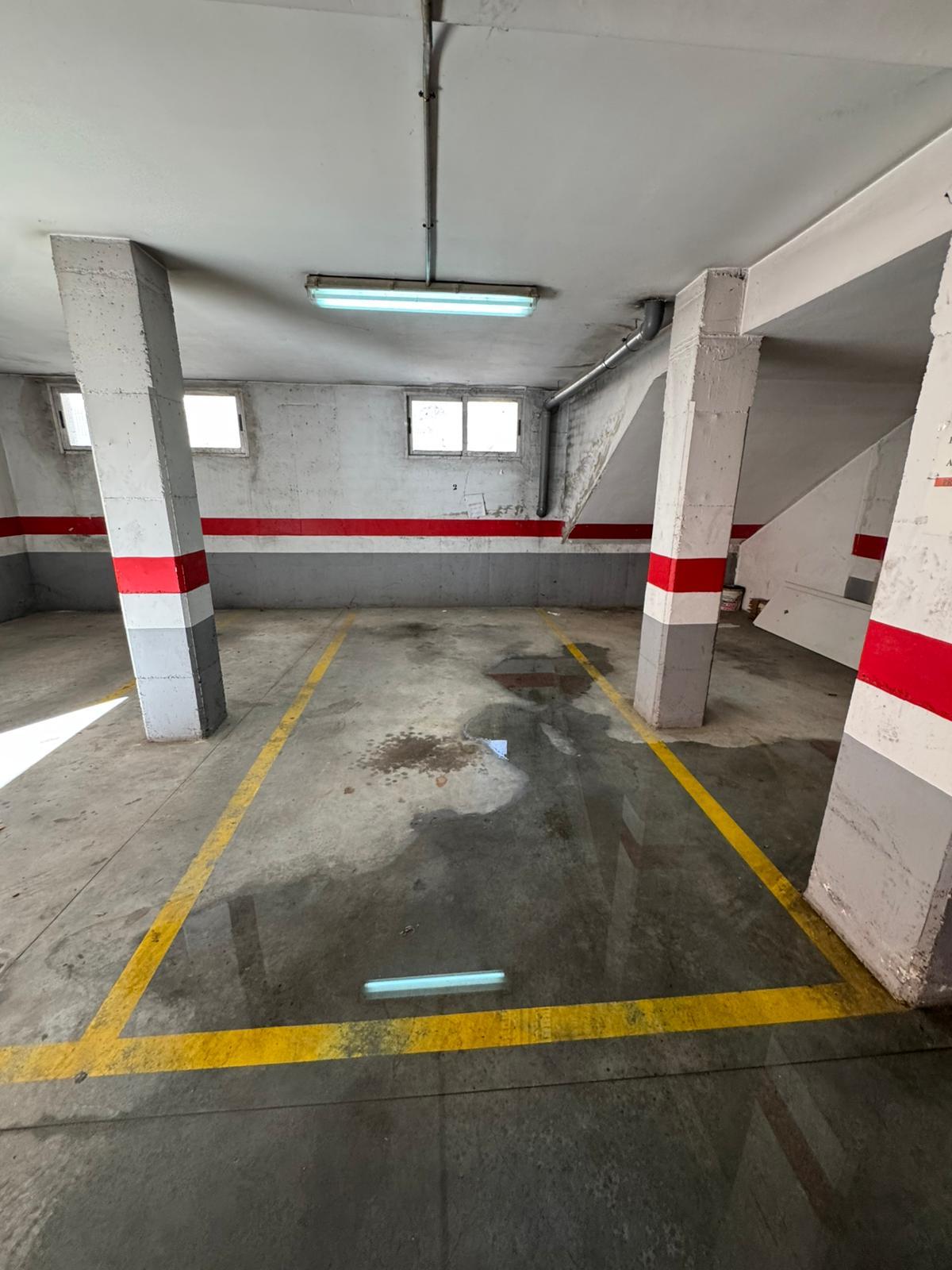Underground parking garage with concrete pillars, yellow parking lines, and wet patches on the floor under a fluorescent ceiling light.