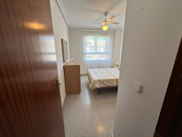 Small bedroom with a double bed, wooden dresser, and a window with sheer white curtains at the far wall. Ceiling fan above bed.