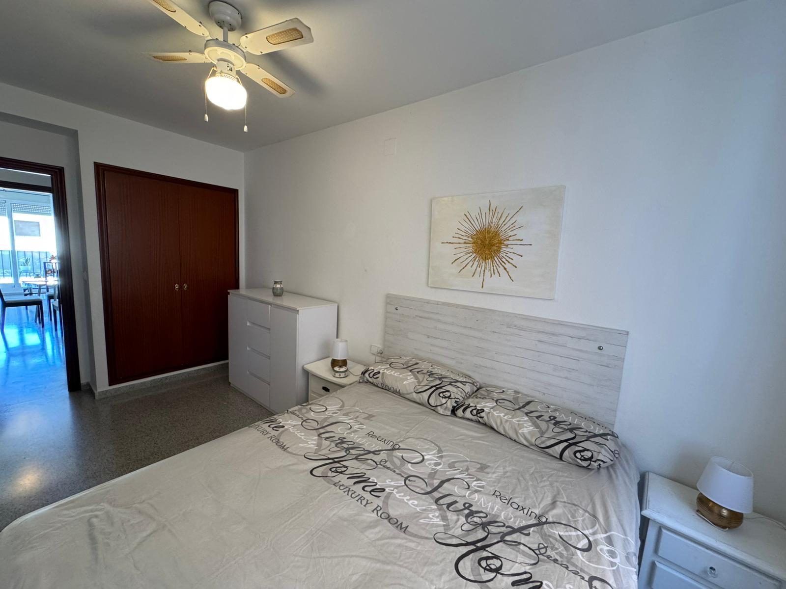 Bright bedroom with a white bed, matching nightstand, and dresser, plus a sunburst wall art above the headboard and a doorway to a dining area outside the room.