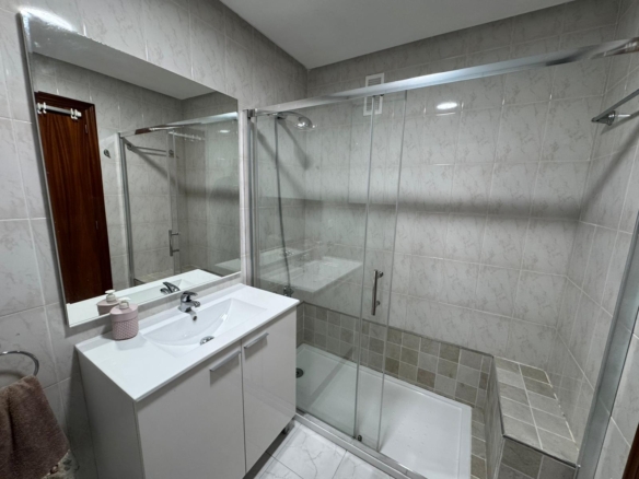 Modern bathroom with a glass shower enclosure, white vanity, and a large wall mirror overlooking beige tiled walls.