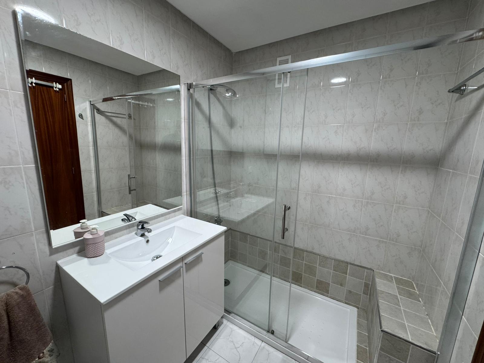 Modern bathroom with a glass shower enclosure, white vanity, and a large wall mirror overlooking beige tiled walls.