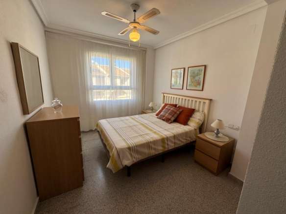 Cozy small bedroom with a wooden bed, striped duvet, two nightstands, dresser, and a ceiling fan with a light. A window with sheer curtains lets in natural light.