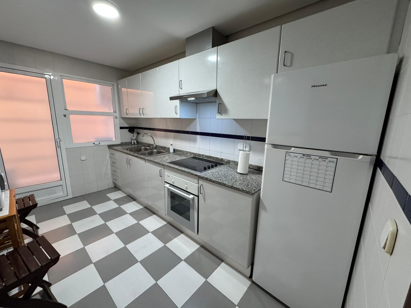 White kitchen with granite countertop, stainless steel sink, and electric stovetop under a range hood, plus a tall white refrigerator on the right.