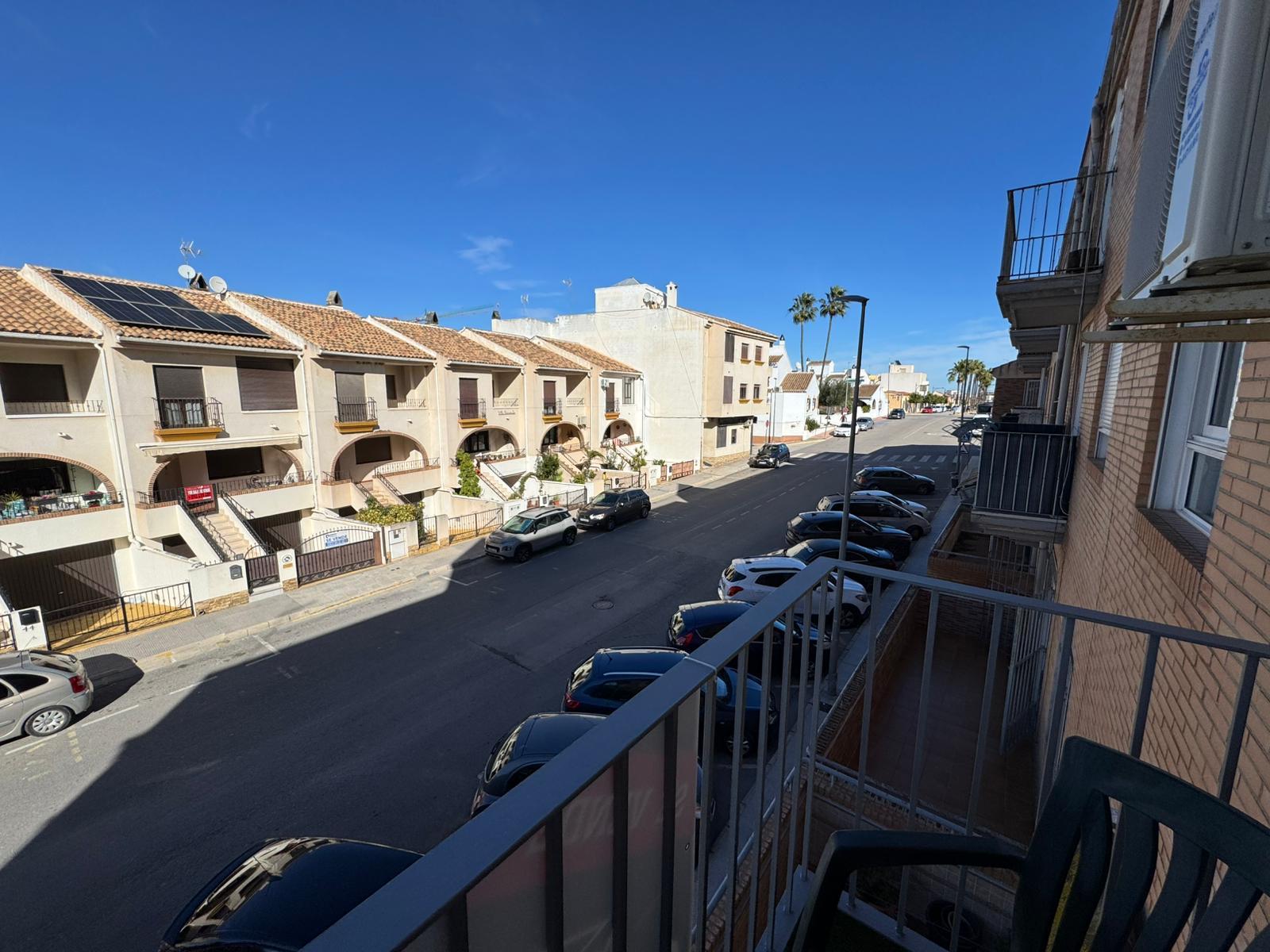 Urban street view from a balcony, beige townhouses with arched entrances and parked cars along the curb under a clear blue sky.