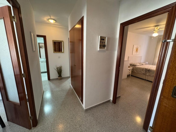 Bright hallway in an apartment with white walls, wooden doorframes, and doors to a bedroom on the right; a potted plant and framed mirror in the hall.