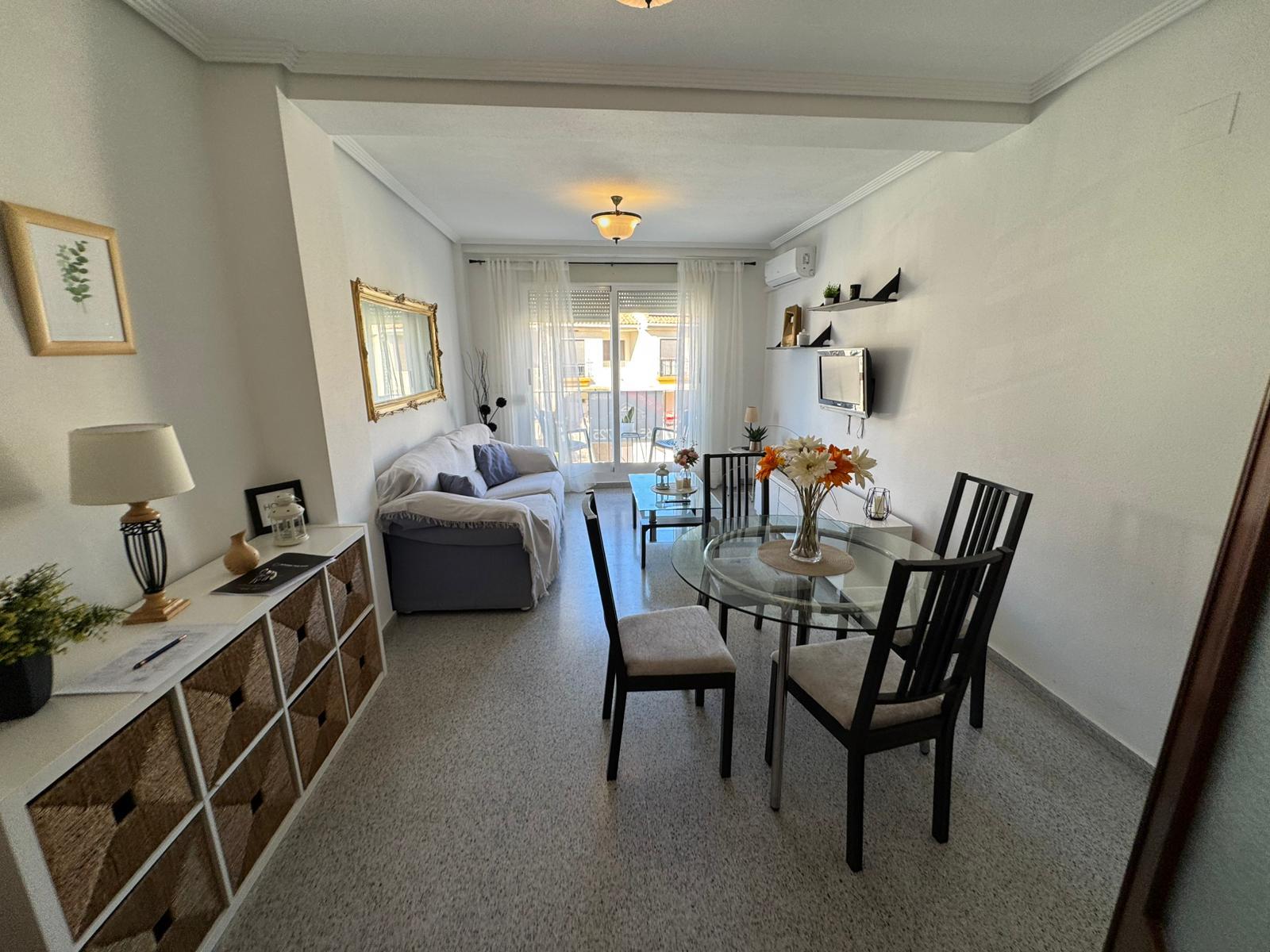 Cozy living room with a glass dining table and six black chairs, a gray sofa facing balcony doors, and sheer white curtains.