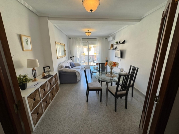Bright living and dining room with a glass round table and four chairs, a gray sofa, white walls, and a balcony with sheer curtains in the background.