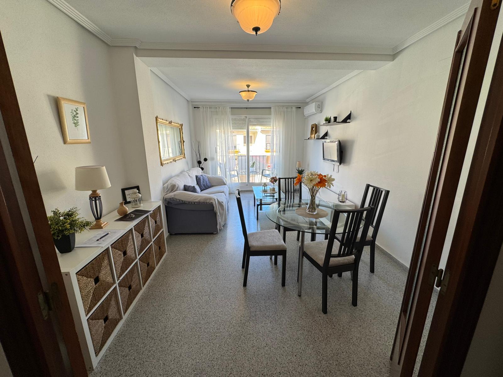 Bright living and dining room with a glass round table and four chairs, a gray sofa, white walls, and a balcony with sheer curtains in the background.