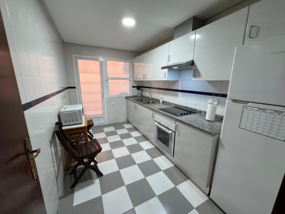 Narrow white galley kitchen with granite countertop, stove, oven, sink, and white cabinets; checkered gray and white floor.