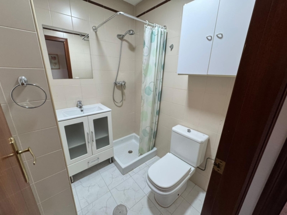 Small tiled bathroom with a white vanity sink, mirrored cabinet, and a shower with a curved curtain rod and light green polka curtain; toilet nearby.