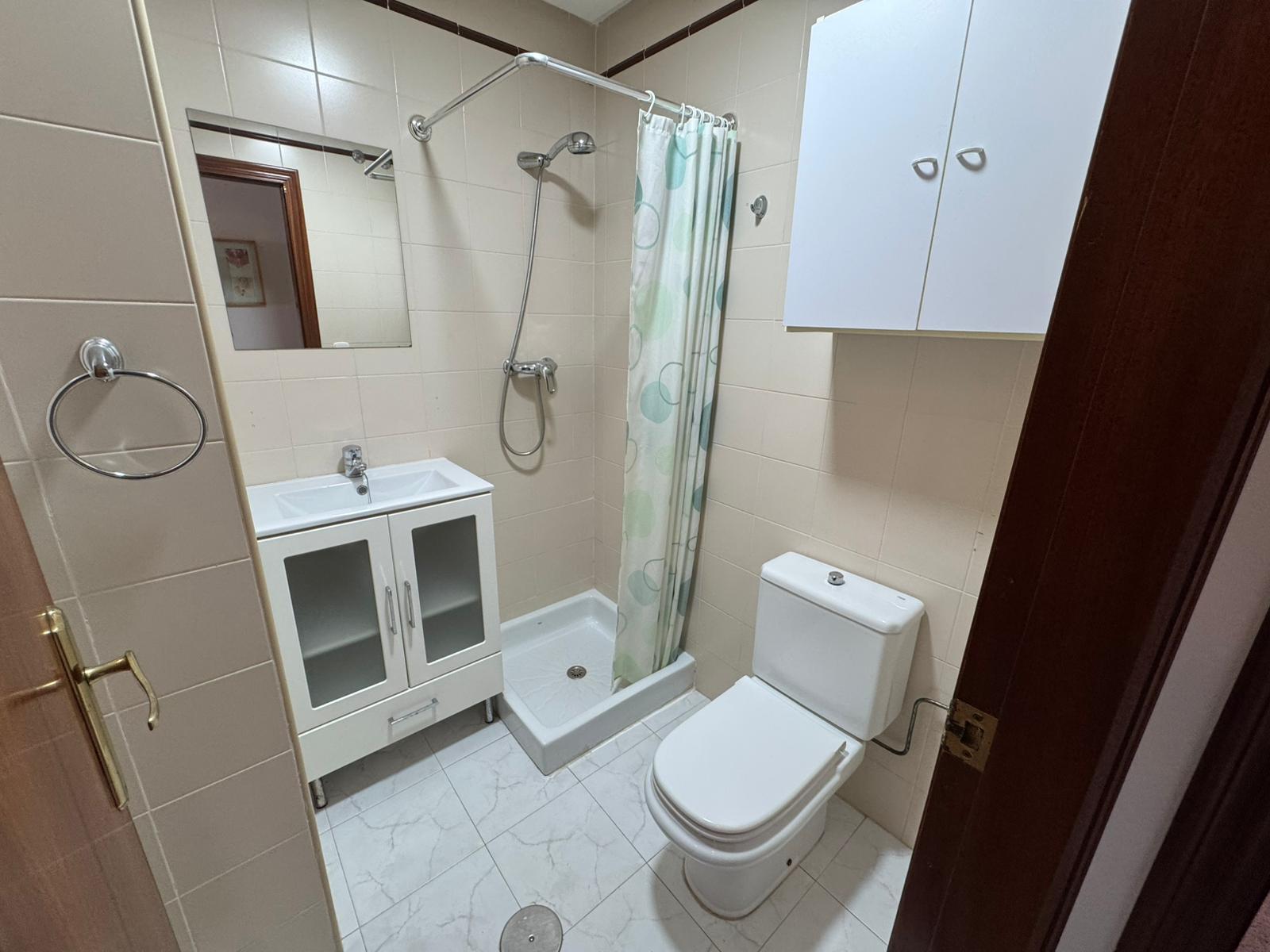 Small tiled bathroom with a white vanity sink, mirrored cabinet, and a shower with a curved curtain rod and light green polka curtain; toilet nearby.