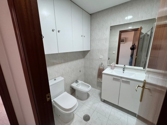 Compact white-tiled bathroom with a wall of cabinets above a toilet and bidet, and a white vanity with mirror.