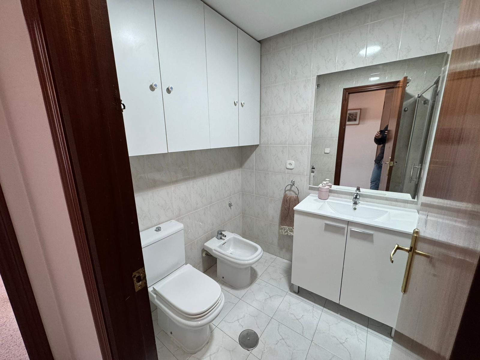 Compact white-tiled bathroom with a wall of cabinets above a toilet and bidet, and a white vanity with mirror.