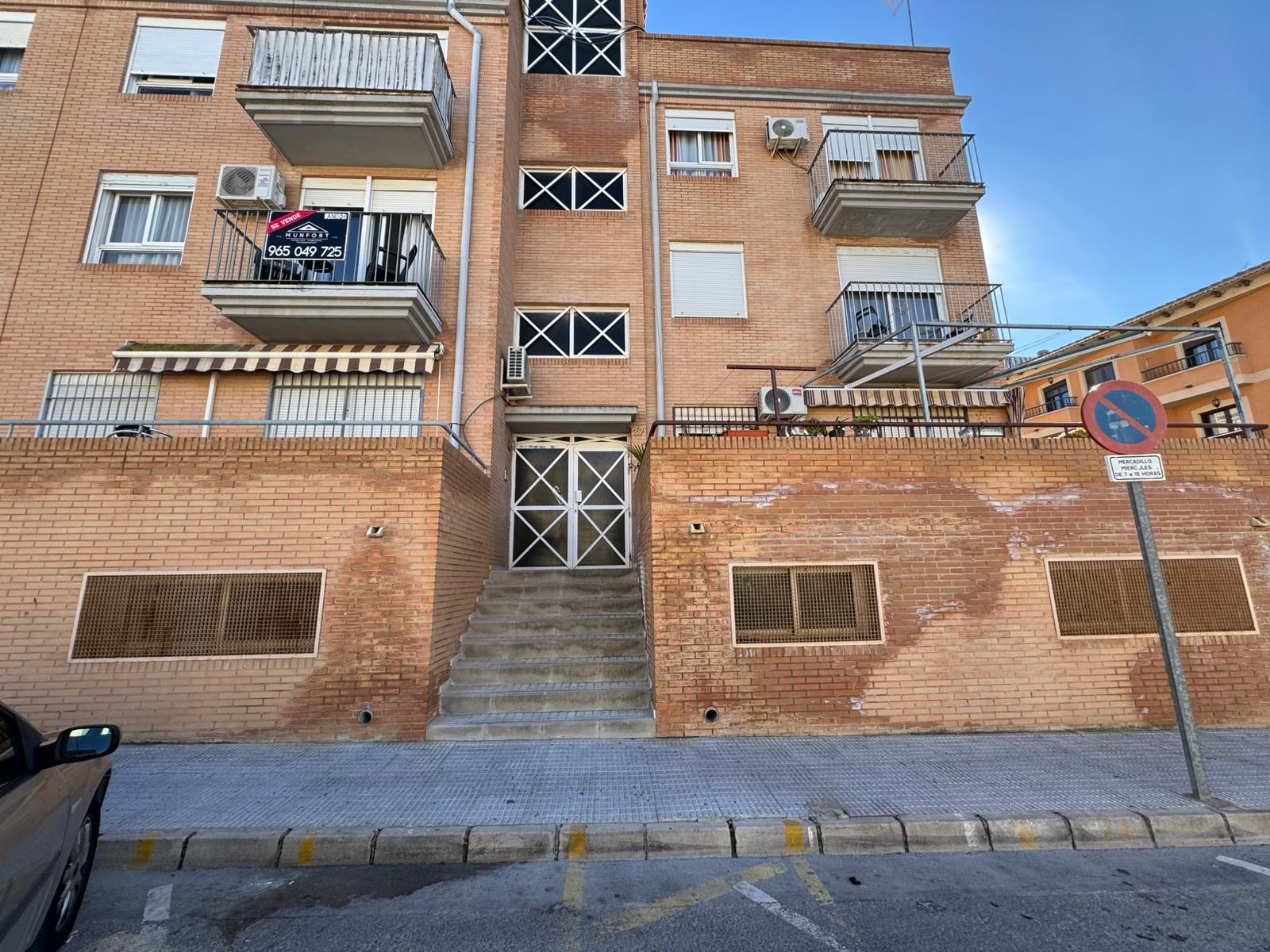 Brick apartment building with central staircase and metal gate at entrance, two balconies on each side, and a street-no-parking sign in foreground.