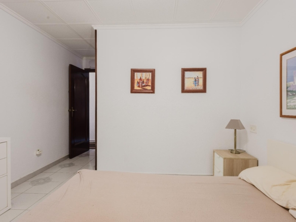 A tidy bedroom with a beige bedspread, white dresser, and a nightstand lamp beside the bed; two small framed pictures on the wall and an open dark wooden door in the adjacent room.