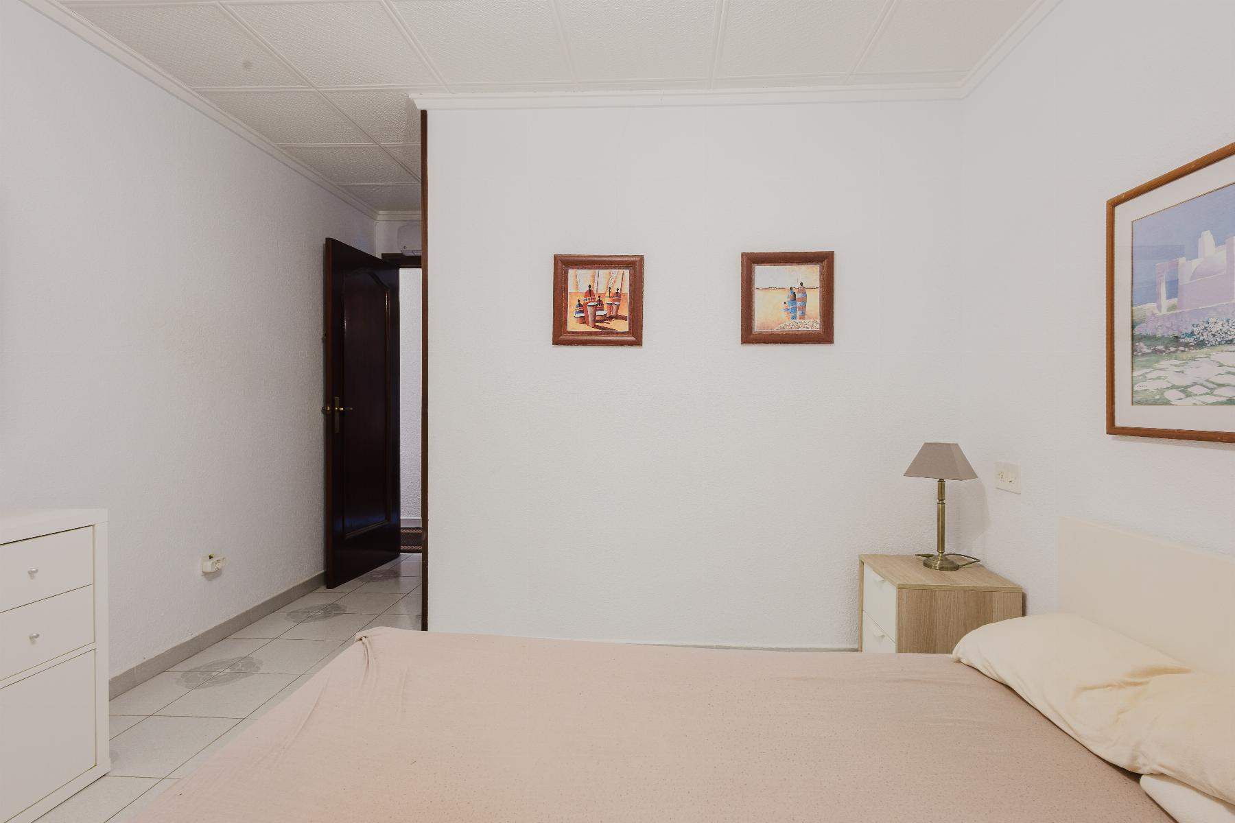 A tidy bedroom with a beige bedspread, white dresser, and a nightstand lamp beside the bed; two small framed pictures on the wall and an open dark wooden door in the adjacent room.