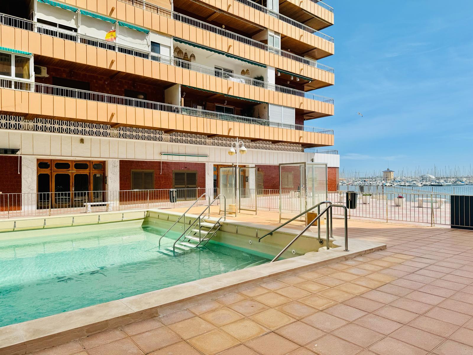 Outdoor swimming pool with metal railings in front of an orange apartment building and a marina in the distance.