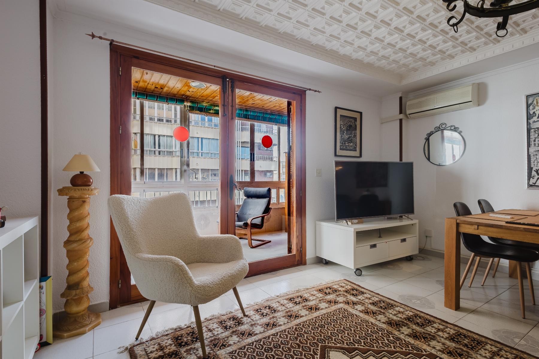 Living room with cream upholstered chairs facing glass doors to a balcony, a patterned rug, and a TV on a white stand.