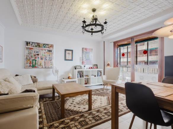 Bright living room with white walls, ornamental ceiling, and a black chandelier; cream sofa, wooden coffee tables, and a patterned rug.