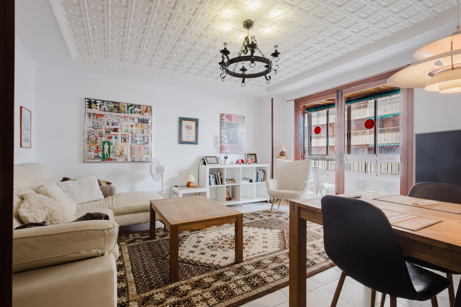 Bright living room with white walls, ornamental ceiling, and a black chandelier; cream sofa, wooden coffee tables, and a patterned rug.