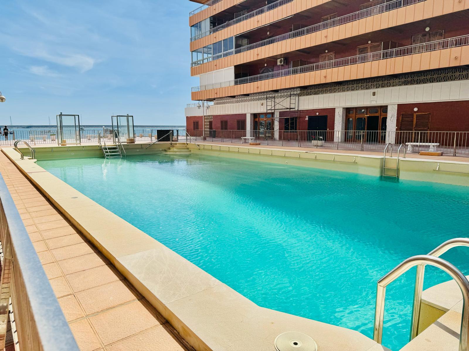 Outdoor hotel pool next to a multi-story orange building, with the ocean and blue sky in the background.