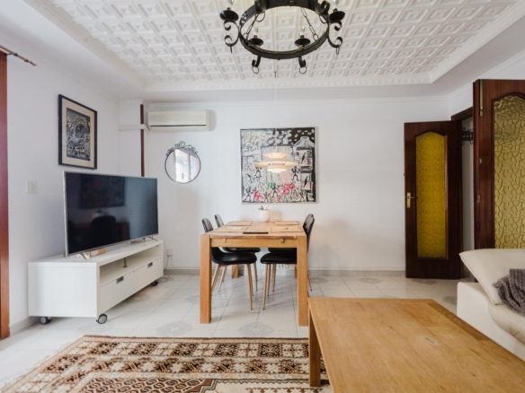 Bright living-dining room with a wooden table and six black chairs, a wall-mounted TV on a white console, and patterned ceiling chandelier.