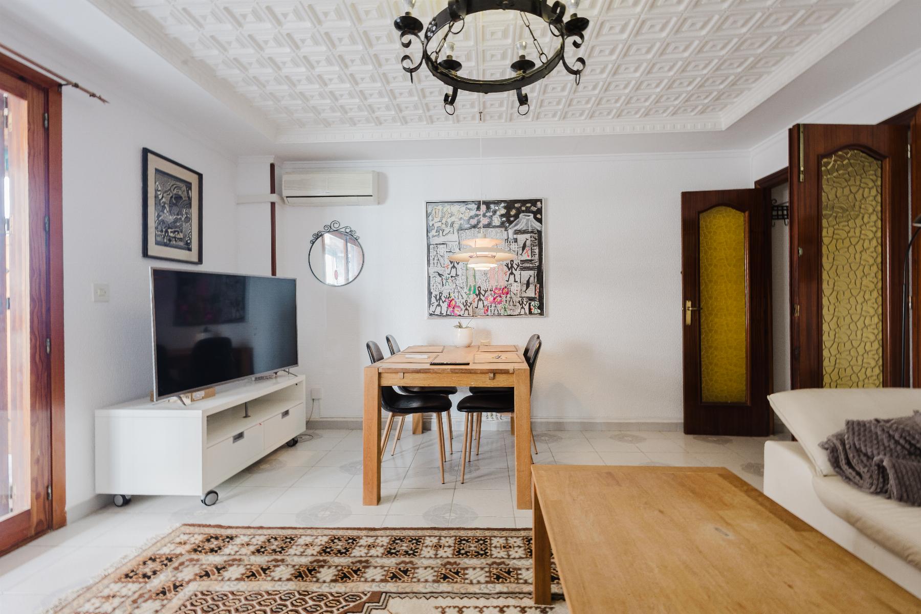 Bright living-dining room with a wooden table and six black chairs, a wall-mounted TV on a white console, and patterned ceiling chandelier.