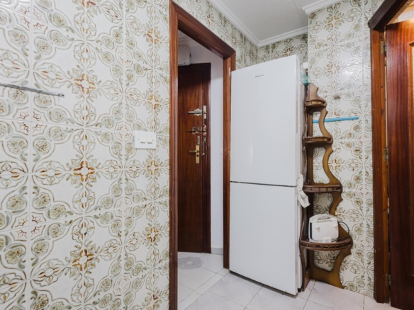 Kitchen corner with patterned tile walls, white fridge, wooden doors, and a curved wooden shelf unit.