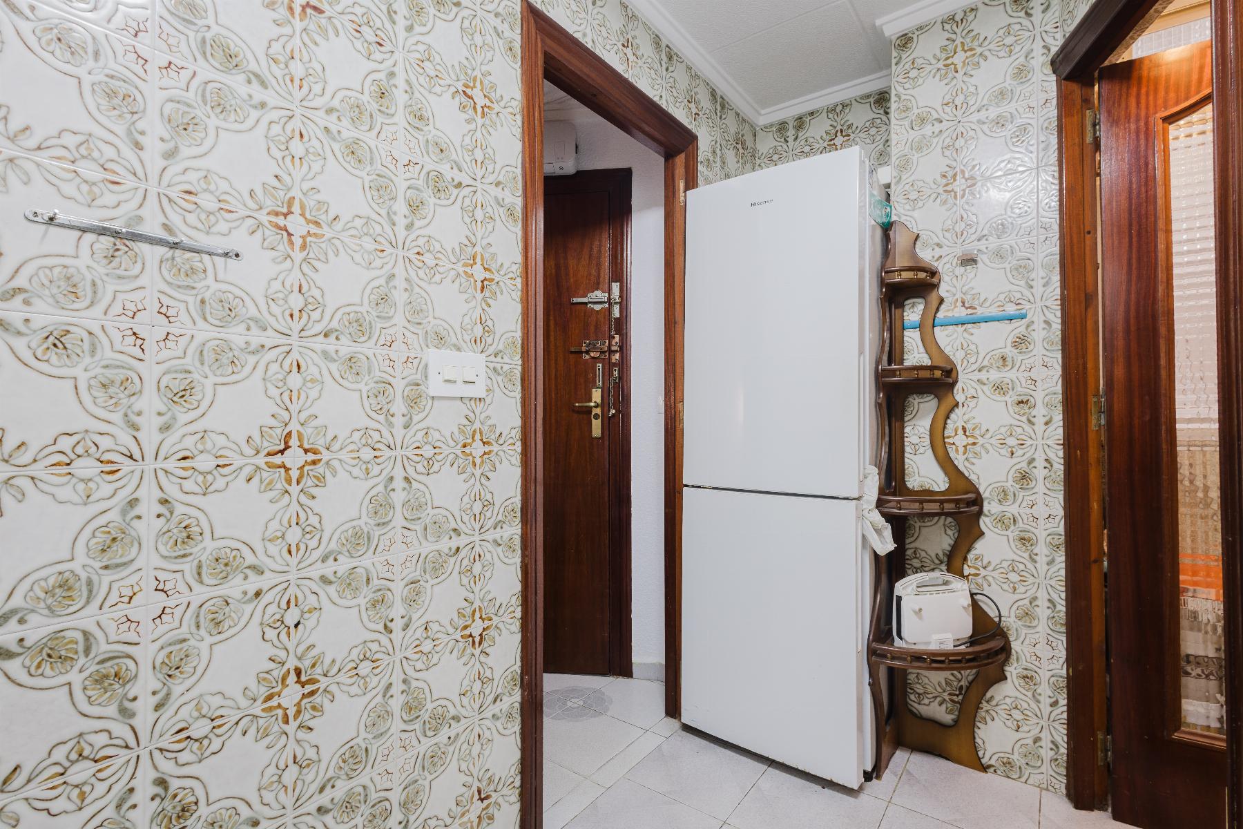 Kitchen corner with patterned tile walls, white fridge, wooden doors, and a curved wooden shelf unit.