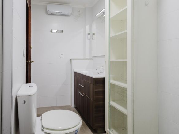 Compact modern bathroom with white tiles, a dark wood vanity, toilet, and glass-front cabinet.