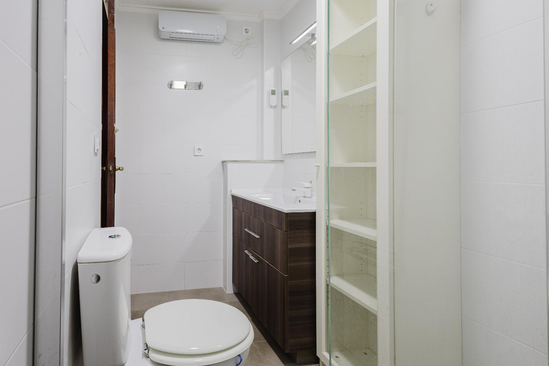 Compact modern bathroom with white tiles, a dark wood vanity, toilet, and glass-front cabinet.
